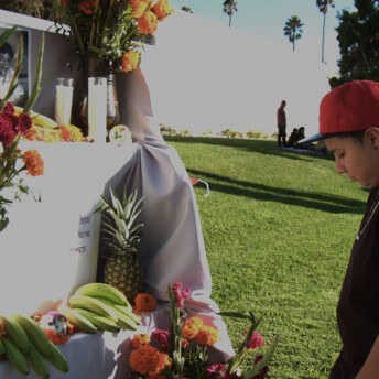 Still from Valentine Road. A young boy stands in front of a memorial for someone. Flowers, candles, and fruit are left behind underneath a portrait of a person.