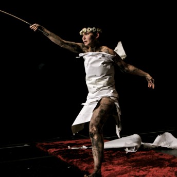 A woman in a white dress with a flower crown is jumping and extending her right arm holding a short sword.