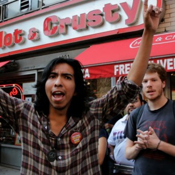 A group of people stands in front of a restaurant. The restaurant sign says "Hot & Crusty". The front-most person in the group holds both of their fists in the air and looks directly at the camera.