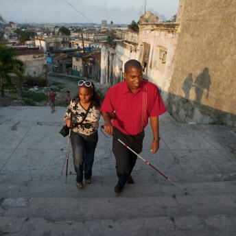 Still from Tocando la Luz. Two persons are walking upstairs, they have their arms intertwined with each other. Each of them uses a cane.