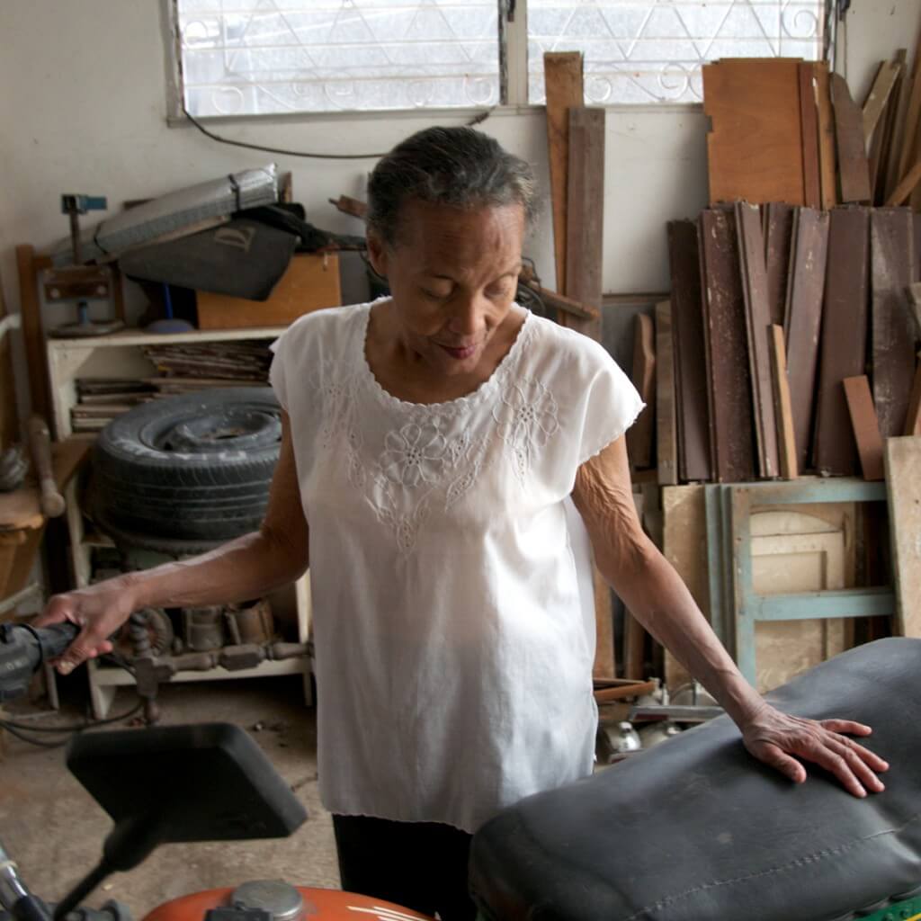 Still from Tocando la Luz. An elderly woman looks down and touches a motorcycle. She is in a room that has wood and a big tire.