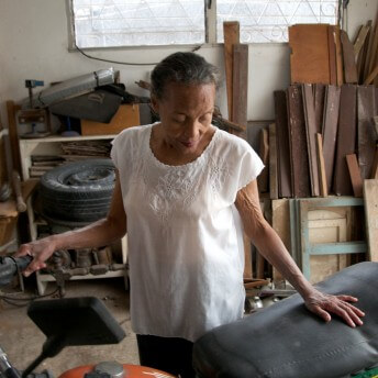 Still from Tocando la Luz. An elderly woman looks down and touches a motorcycle. She is in a room that has wood and a big tire.