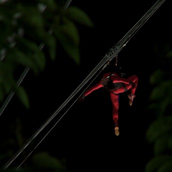 In the night, a human figure wearing a red body suit is hanging from a cable.