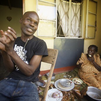 A man is sitting on a chair, talking to someone who is out of frame. Behind him, there is a woman sitting on the floor having lunch, there are dishes on the ground.