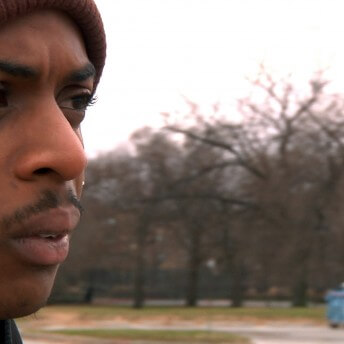A still from The Homestretch. A close-up shot of a person's face. They have a stern look and look off to the right of the camera. They are wearing a maroon beanie and standing in a field on a cloudy day.