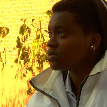 A still from The Homestretch. A close-up shot of a person in a white jacket and maroon beanie outside, in front of a brightly lit wall in the background. The wall appears yellow from the bright sun.