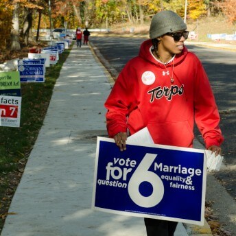 Still from The New Black. A person wearing a hat, sunglasses, and a bright red sweatshirt walks down a sidewalk. They are holding a sign that reads: "Vote for Question 6: Marriage Equality & Fairness".