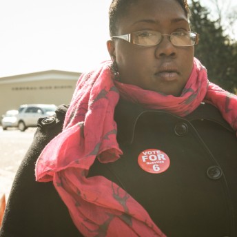 Still from The New Black. A person stands facing the camera and looks directly into the lens. They are wearing a black button-down jacket and a red scarf. They also have a red pin that reads "Vote For Question 6".