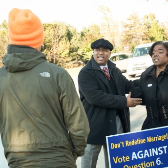 Still from The New Black. Two people stand behind a sign that reads, "Don't redefine marriage! Vote Against Question 6". Another person stands in front of the sign, facing the two people behind it. They are wearing an orange hat and green jacket, their back is to the camera.