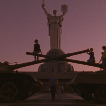 Still from The Vanquishing of the Witch Baba Yaga. Two army tanks face each other with children atop. A large statue stands tall in the background. Color photograph.