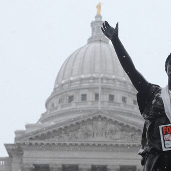 A statue of a woman in front of a governmental building has a hanging sign that says "For sale. Wisconsin. Call Scott Walker"