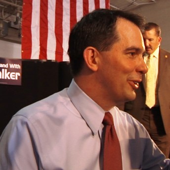 A man wearing a shirt and a tie is greeting people out of frame. In the back, there are posters that say "We Stand with Walker."