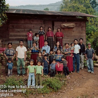 Still from Granito. Outdoor group photo of about 25 people–including, men, women, and children. The women wear patterned traditional Guatemalan indigenous clothing and headbands. All people are staring straight ahead, unsmiling. Photo credit, Dana Lixenberg, is listed on the bottom left of the image.