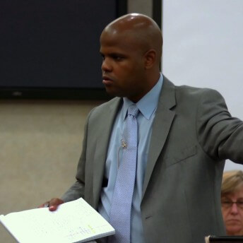 Still from Gideon's Army. A man in a grey suit jacket and light blue dress shirt and tie is pointing with his hand to his left. He is holding a notepad in his right hand.