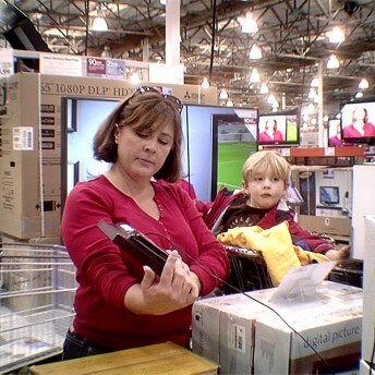 Still from Xmas Without China. A woman stands in an electronics store, holding an electronic with a long cord in one hand. She wears a red top, sunglasses on top of her head, and has short dark hair. A young child sits in a cart behind her. He wears a coat and has blonde hair.