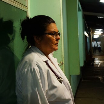 Still from A Quiet Inquisition. Profile shot of a doctor wearing a robe, her hair is in a bun, and she wears thick glasses. She is standing against a green wall.