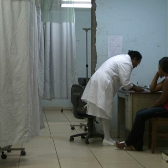 Still of A Quiet Inquisition. Full shot of a nurse leaning on a desk to write on a paper while a woman patient is waiting next to her sitting on a chair.