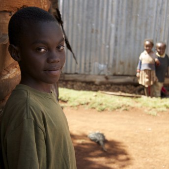 Still from A Small Act . Medium profile shot of a boy with an olive t-shirt looking at the camera, in the back and out of focus there are other two children.
