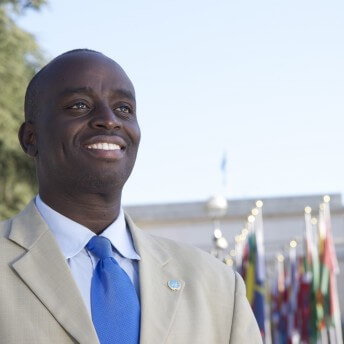 Still from A Small Act. Low angle medium shot of a man looking up, wearing a white suit and blue tie, in the background there are out-of-focus flags on flag poles.