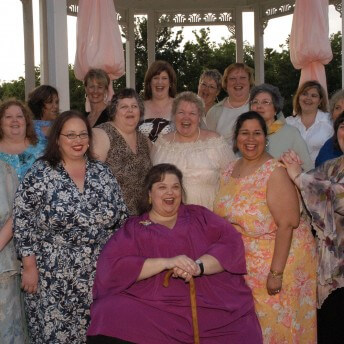 Still from All of Me. Group photo of sixteen women with dresses smiling at the camera.