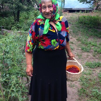 A photo of an older woman wearing a long black skirt and a colorful patterned top. She wears a headscarf and holds a basket of berries.
