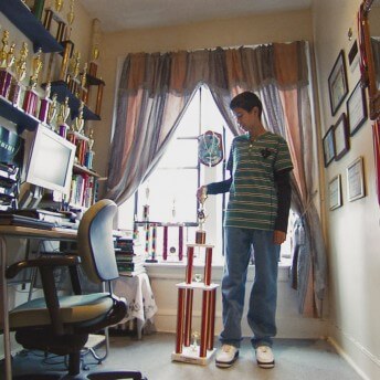 A teenager is standing in a room full of trophies, he has a trophy next to him and is looking at it.