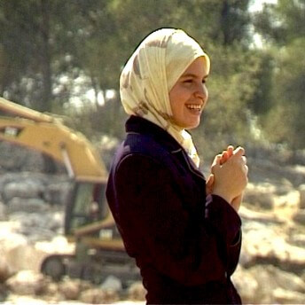 A woman is standing on a construction site, she is wearing a yellow hijad and holding her hands together in front of her chest.