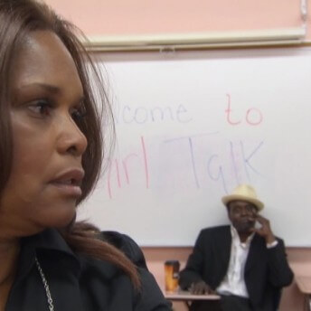 Close up to a woman's face. Behind her there is a whiteboard and a man sitting on a chair.