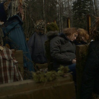 Still from LoveTrue. Two young folks are sitting and reclining their heads on each other, they wear winter jackets. They are surrounded by child-like manequins.