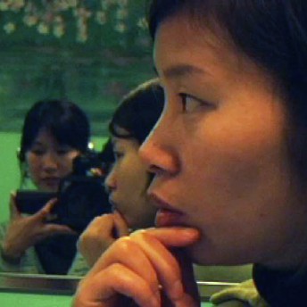In the foreground a woman is looking away from the camera, her hand is touching her chin. In the background, there is a mirror reflection of Filmmaker Debbie Lum recording with a hand-held camera.