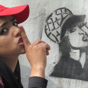 Sonita imitates a graffiti painted on a wall, she holds her index finger in front of her mouth and looks at the camera with her head tilted to the back.