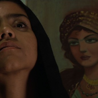 Still from Sonita. Sonita in front of the camera tilts her chin up, behind her there is a mural of a woman.