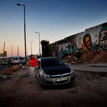 Still from Speed Sisters. A car in a street with no pavement, in front of a colorful mural. There is a person with half of their body out of the co-pilot window and wearing a bright red jacket.