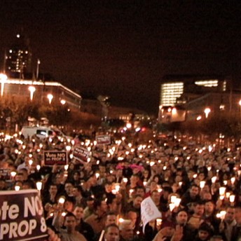 A still from The Campaign. A wide shot of a large group of people gathered during the night time. They are holding a vigil, many people hold small lights. Some signs are visible, one reads "Vote NO PROP 8".