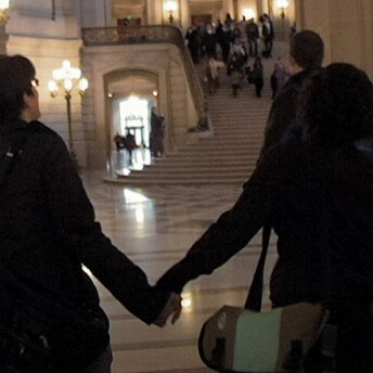 A still from The Campaign. Two people hold hands and walk through a large hall. A grand staircase sits behind them, in the distance with a group of people gathered at the top.