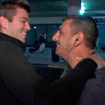 Still from The List. Two people stand facing one another. The person on the left has their hand on the other person's shoulder. They smile at one another.