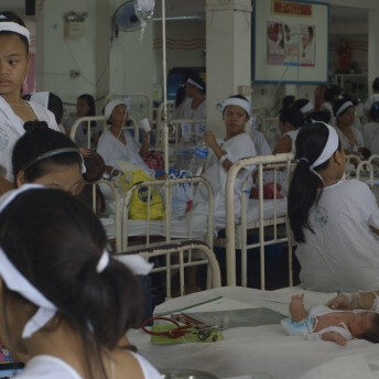 Still from Motherland. A hospital ward with many beds filled with women in various positions–standing, sitting, and talking with one another. In the center, an infant is lying on an empty bed with women surrounding them.