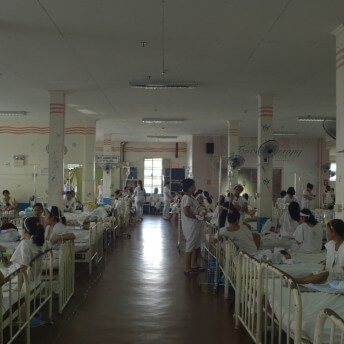 Still from Motherland. A hospital ward with many beds filled with women in various postions - standing, sitting, and talking with one another.