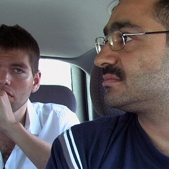 Still from The List. Two people sit inside a car, one person sits in the front seat, and the other in the seat behind. The person in the front looks to their right, away from the camera. The person in the back seat looks forward, their eyes resting slightly above the camera lens.