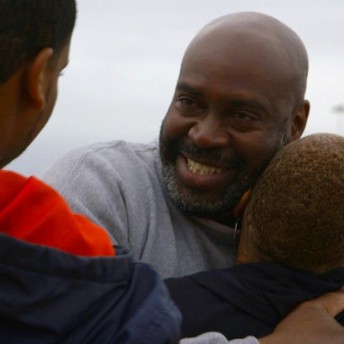 Still from The Return. Two people hugging one another. The person in the front faces away from the camera. The person behind faces the camera and is smiling. They look at the third person on the left side of the frame, who also faces away from the camera.
