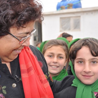 A few young girls wearing head coverings smile at the camera. An adult standing beside them smiles down at them. Color photograph.