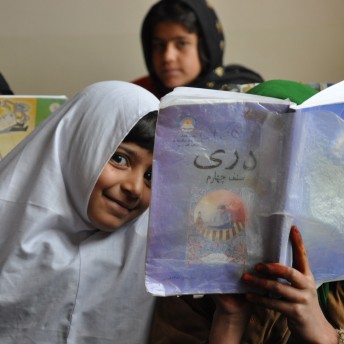 A young girl wearing a head covering looks playfully at the camera. Beside her is someone holding up a book to their face. Another girl who is out of focus looks at the camera in the background. Color photograph.
