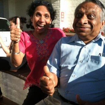 Still from Meet the Patels. A man in a blue shirt and woman in a pink shirt, looking directly at the camera, giviving an enthusiastic thumbs up. There is another woman as well as a car in the background.
