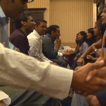 Still from Meet the Patels. There is a row of men on the left sitting in front of a row of women on the right. A couple in the foreground are shaking hands.