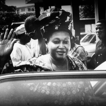 A person wearing patterned clothing and head scarf with their hand up standing behind a car door. There are people and cars in the background. Black and White photograph.