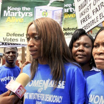 A person speaking into a microphone. People are surrounding this person wearing the same t-shirt holding signs that say keep the martyrs alive. Color photograph.
