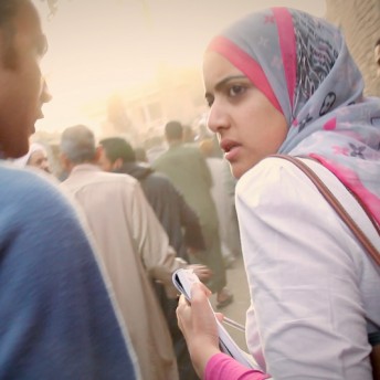 Still from Words of Witness. A woman in a white top and a gray, pink, and black hijab looks over her shoulder. A man stands beside her wearing a blue shirt.