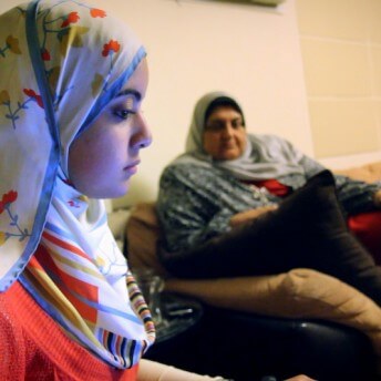 Still from Words of Witness. Two women sit on a sofa. One wears a white top and a white hijab with red flowers. The other wears a gray hijab and a top.