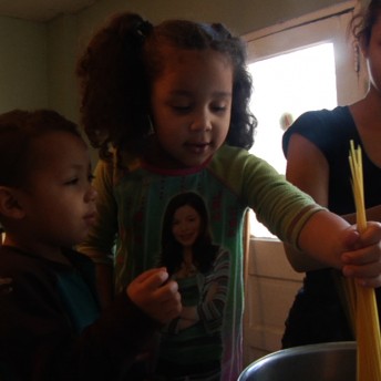 Still of A Place at the Table. A young woman is cooking spaghetti with a child and a toddler; they are putting the pasta in a pan.