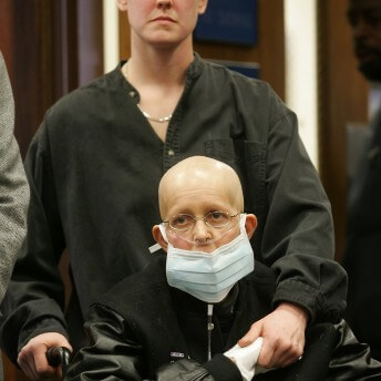 Still from Freeheld. A woman in a black letterman jacket is sitting in a wheechair. She is bald, wears silver-rimmed glasses, a nasal breathing tube, a mask over her mouth, and white gloves. She holds the hand of a short-haired woman who is standing behind her and is holding onto her wheelchair. Both women are looking at a point off-screen.